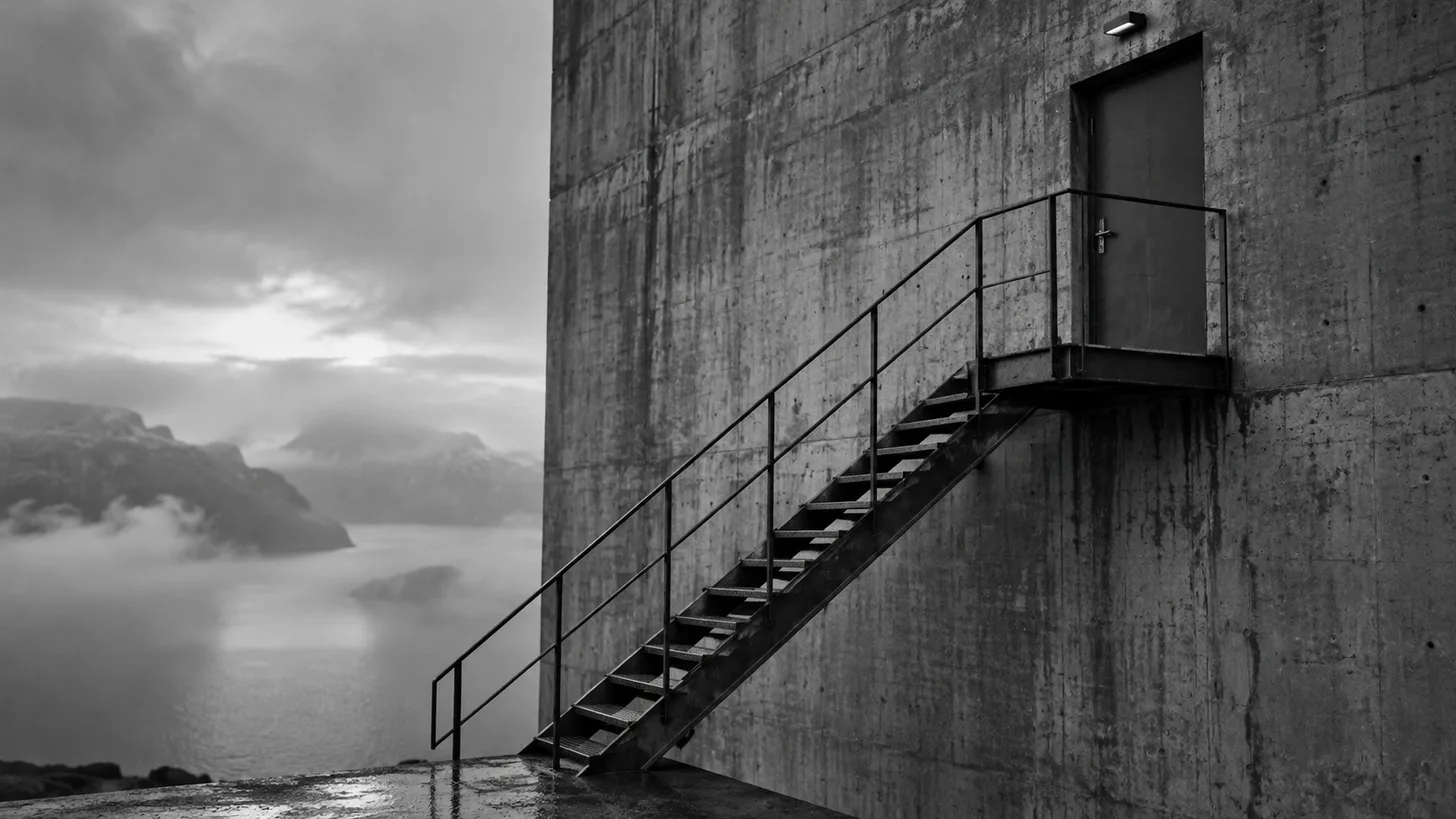 A minimal metal staircase ending at a locked door high on a stark concrete wall.