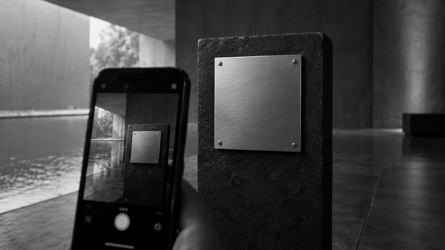 A smartphone camera pointed toward a blank square metal plaque on a dark stand,